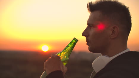Close-up-of-a-man-drinking-beer-at-sunset-standing-on-the-roof-of-a-building-against-the-background-of-a-beautiful-evening-city.